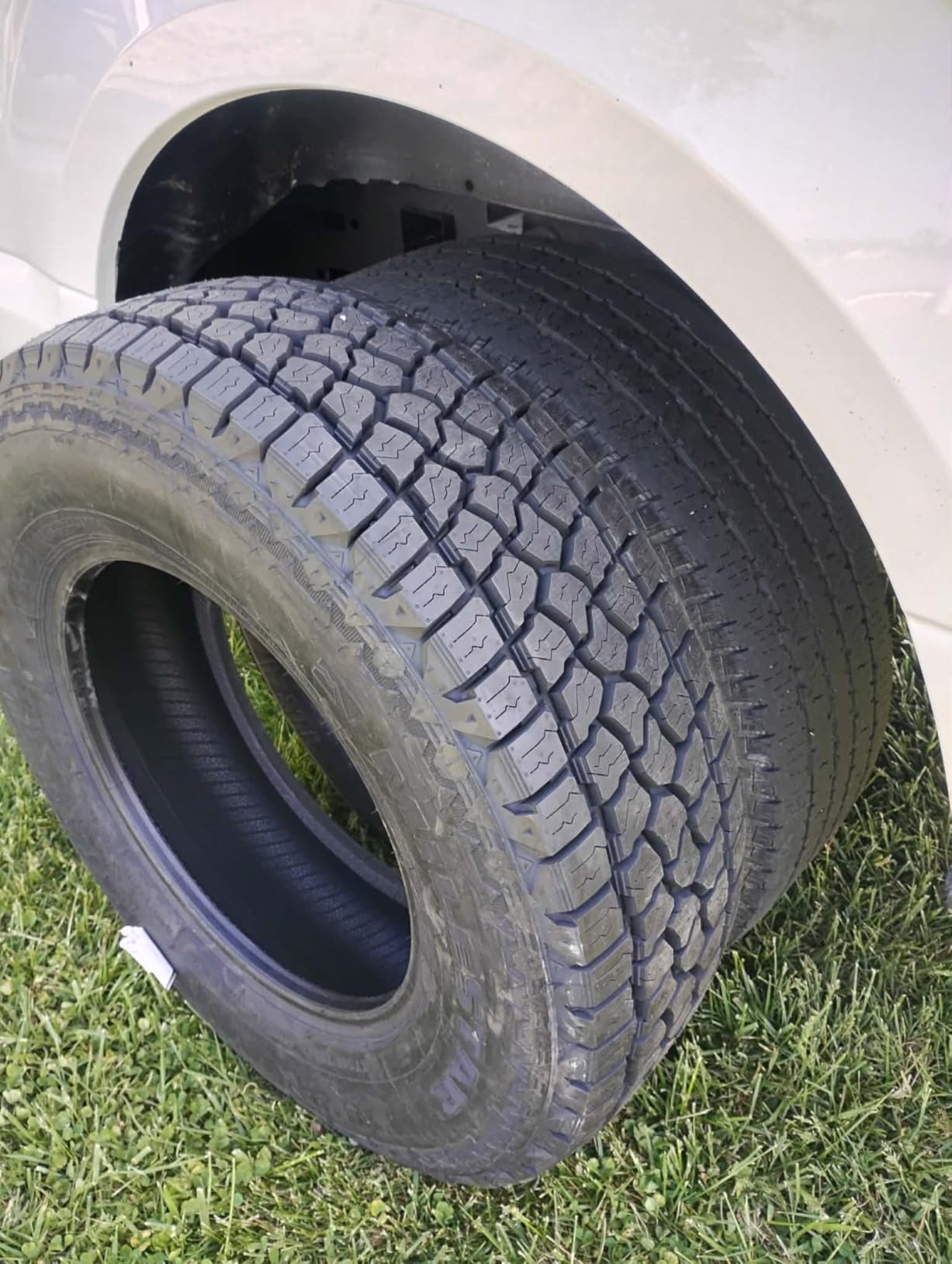 Milestar Patagonia A/T tire stood against the front wheel arch of a white truck on grass, tread blocks and shoulder lugs in full sun