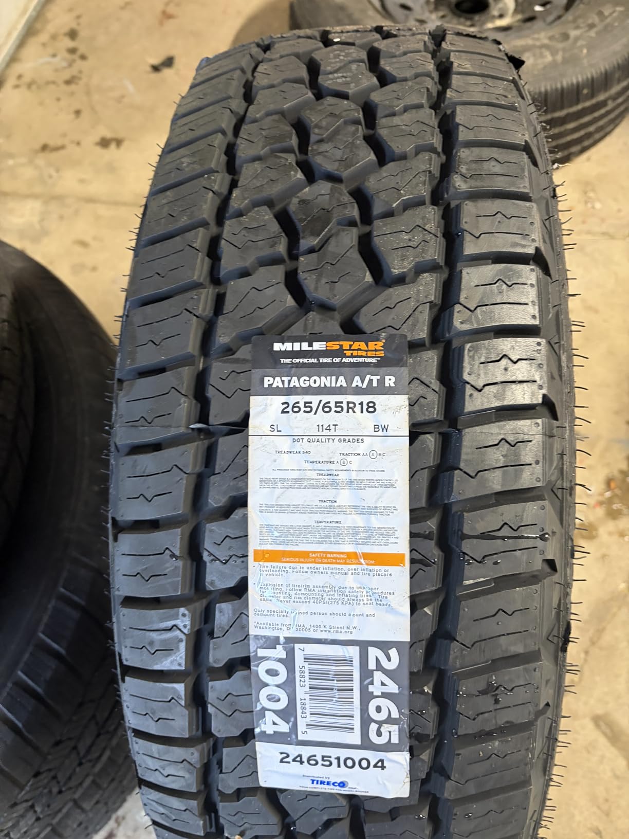 New Milestar Patagonia A/T R in 265/65R18 lying tread up on a shop floor, spec label reading SL 114T BW still in place