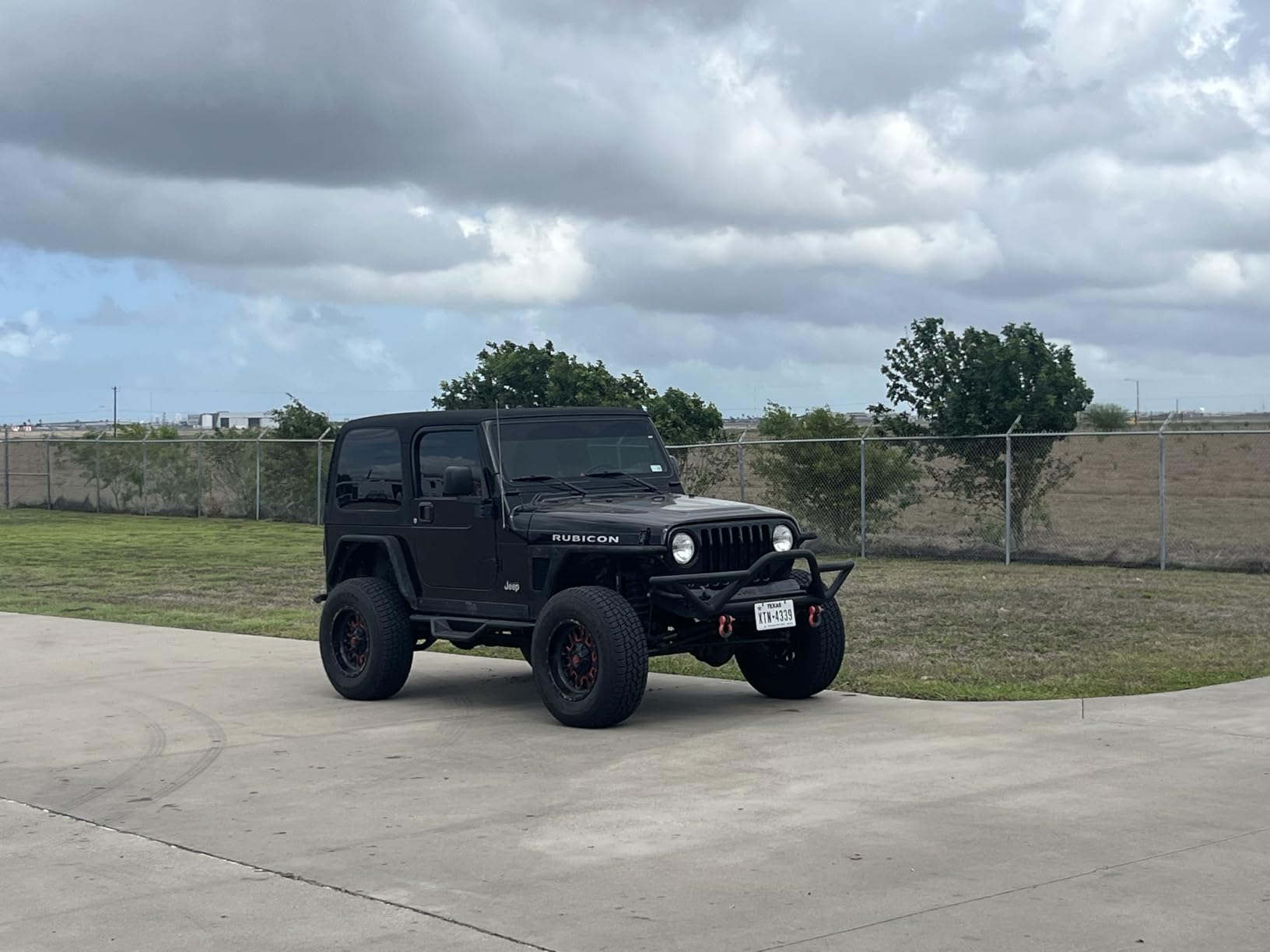 Black Jeep Wrangler Rubicon on Patagonia tires standing on a concrete pad, chain-link fence and open field behind it